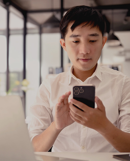 How Gen Z is reshaping investing behaviour and wealth management A young professional seated at a desk looks at a smartphone while a laptop is open in front of them, illustrating digital-first engagement alongside traditional work tools.