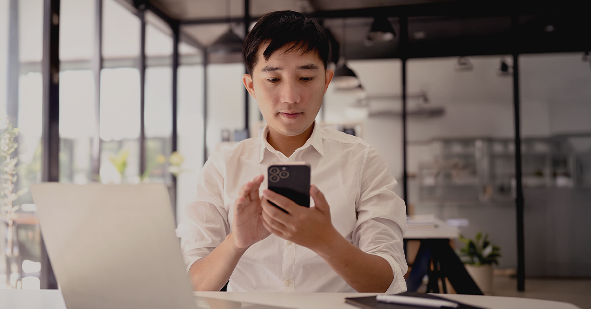 How Gen Z is reshaping investing behaviour and wealth management A young professional seated at a desk looks at a smartphone while a laptop is open in front of them, illustrating digital-first engagement alongside traditional work tools.